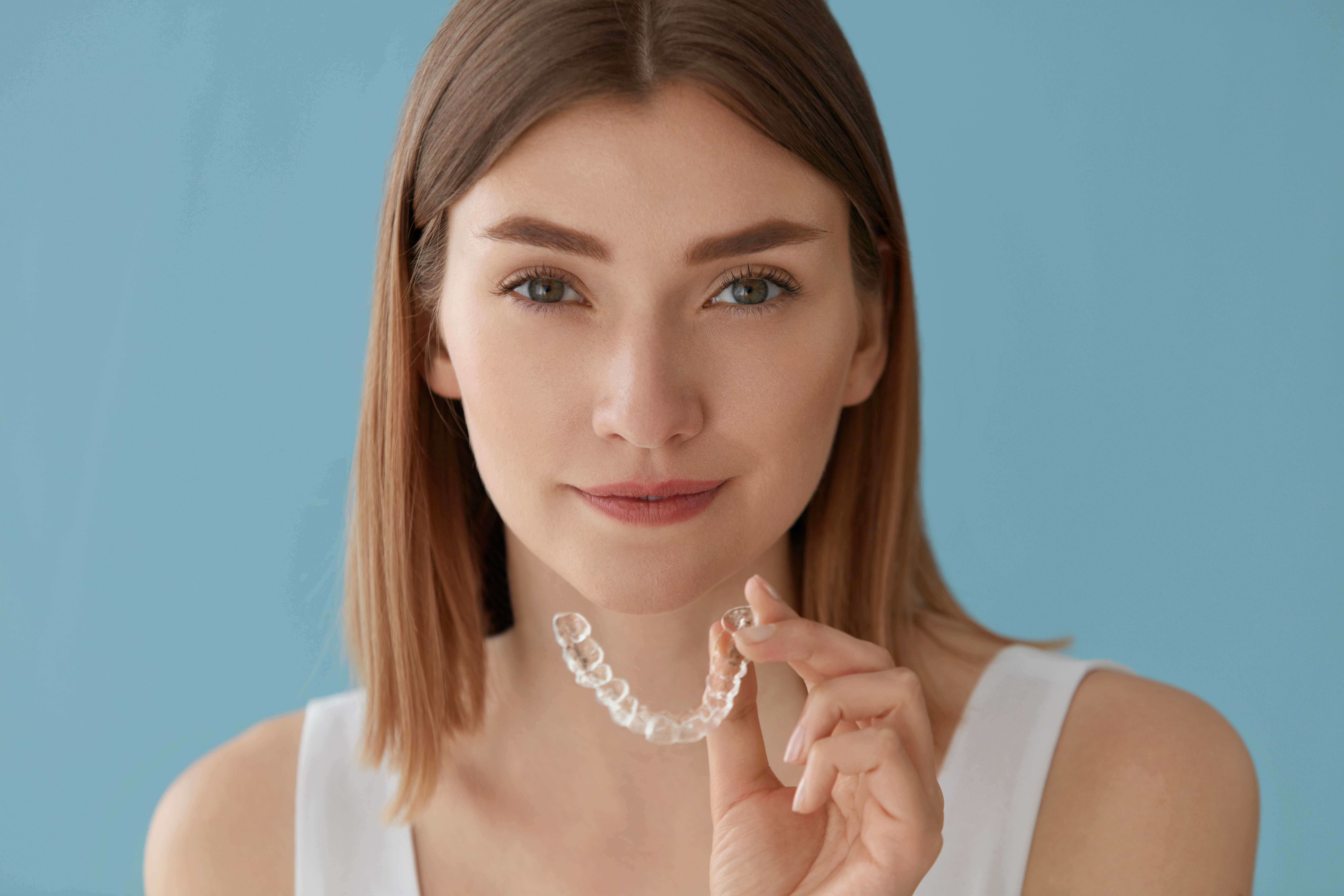 Young woman holding clear aligner against a light blue background, showcasing discreet orthodontic treatment option for straighter teeth.