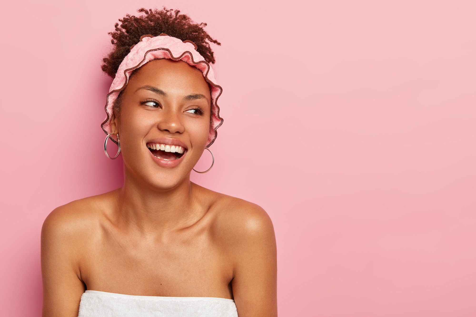 Smiling woman with curly hair wearing a pink headband and hoop earrings, against a pink background, representing confidence and beauty.