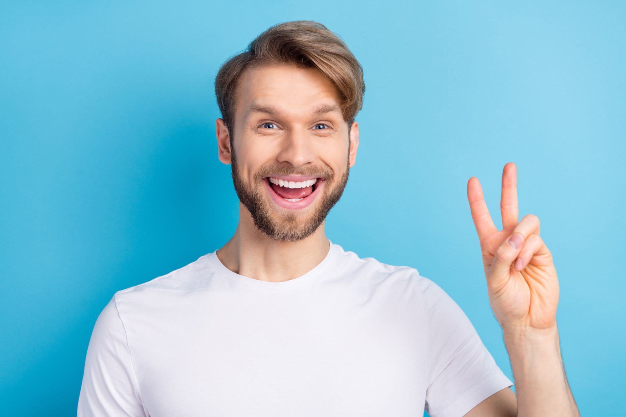 Smiling man in a white t-shirt showing a peace sign against a blue background, representing confidence and satisfaction with clear aligners from Bond Orthodontics in Northbrook, IL.